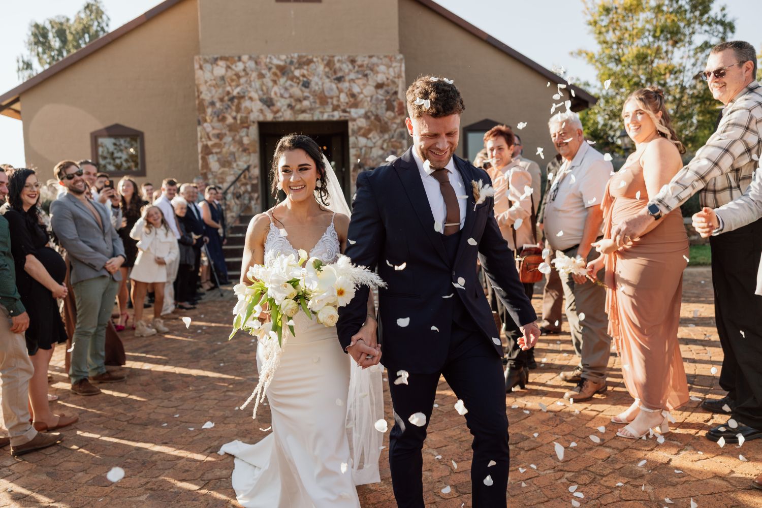 Wedding couple walking out of chapel smiling while guests throw white flower petals in celebration